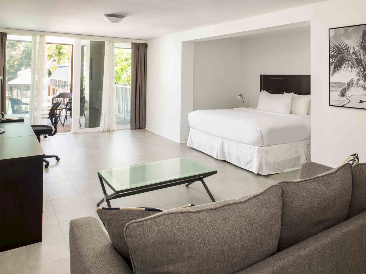 The image shows a modern hotel room with a bed, sofa, desk, TV, coffee table, and a balcony, decorated with a palm tree wall art.