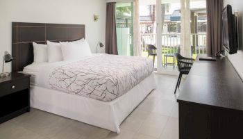 The image shows a tidy hotel room with a king-sized bed, bedside tables, a desk, a flat-screen TV, and a balcony with chairs.