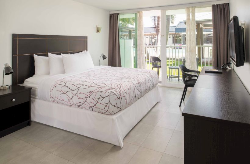 The image shows a tidy hotel room with a king-sized bed, bedside tables, a desk, a flat-screen TV, and a balcony with chairs.