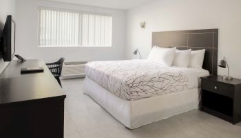 A modern bedroom with a bed, nightstands, TV, and a window with blinds. The room is minimalist with a neutral color palette and simple decor.