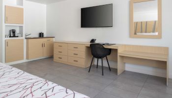 This image shows a modern hotel room with a TV, desk, chair, mirror, cabinets, and a coffee maker on a counter, and bed in the foreground.