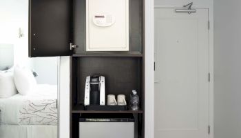 A hotel room with an open closet containing a safe, coffee maker, cups, and water bottles. A bed and entry door are also visible in the room.