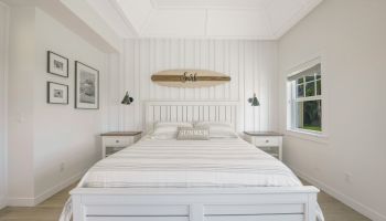 A bedroom with a white bed, striped bedding, surfboard decor, wall art, bedside tables with lamps, and a window letting in natural light.