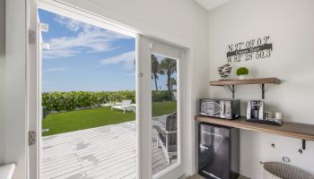 A room with a small kitchenette featuring a microwave, coffee maker, mini fridge, and a view of a deck with lounge chairs and green foliage outside.