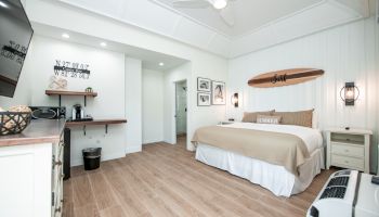 A modern bedroom with a king bed, neutral decor, wall art with a surfboard, and a TV. There’s also a small workspace and coffee station.