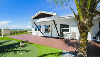 A modern house with a patio, a small putting green, two white chairs, a palm tree in the foreground, and a clear blue sky in the background.