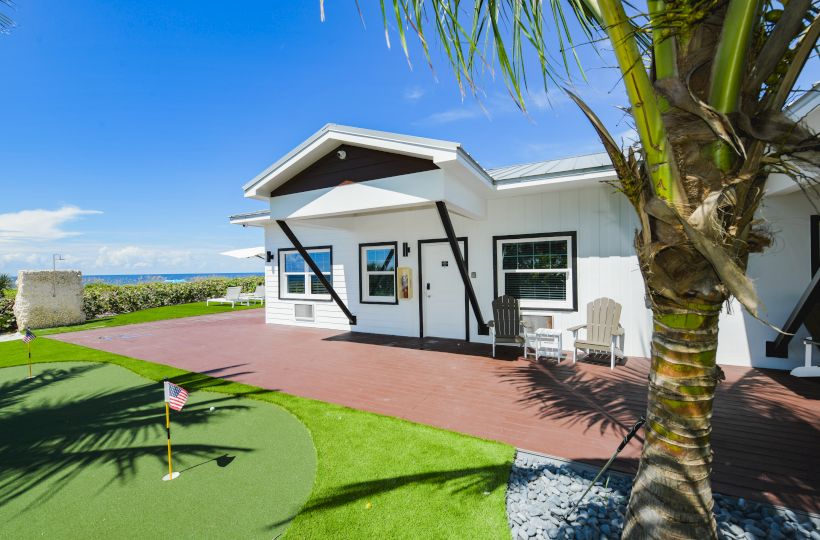 A modern house with a patio, a small putting green, two white chairs, a palm tree in the foreground, and a clear blue sky in the background.