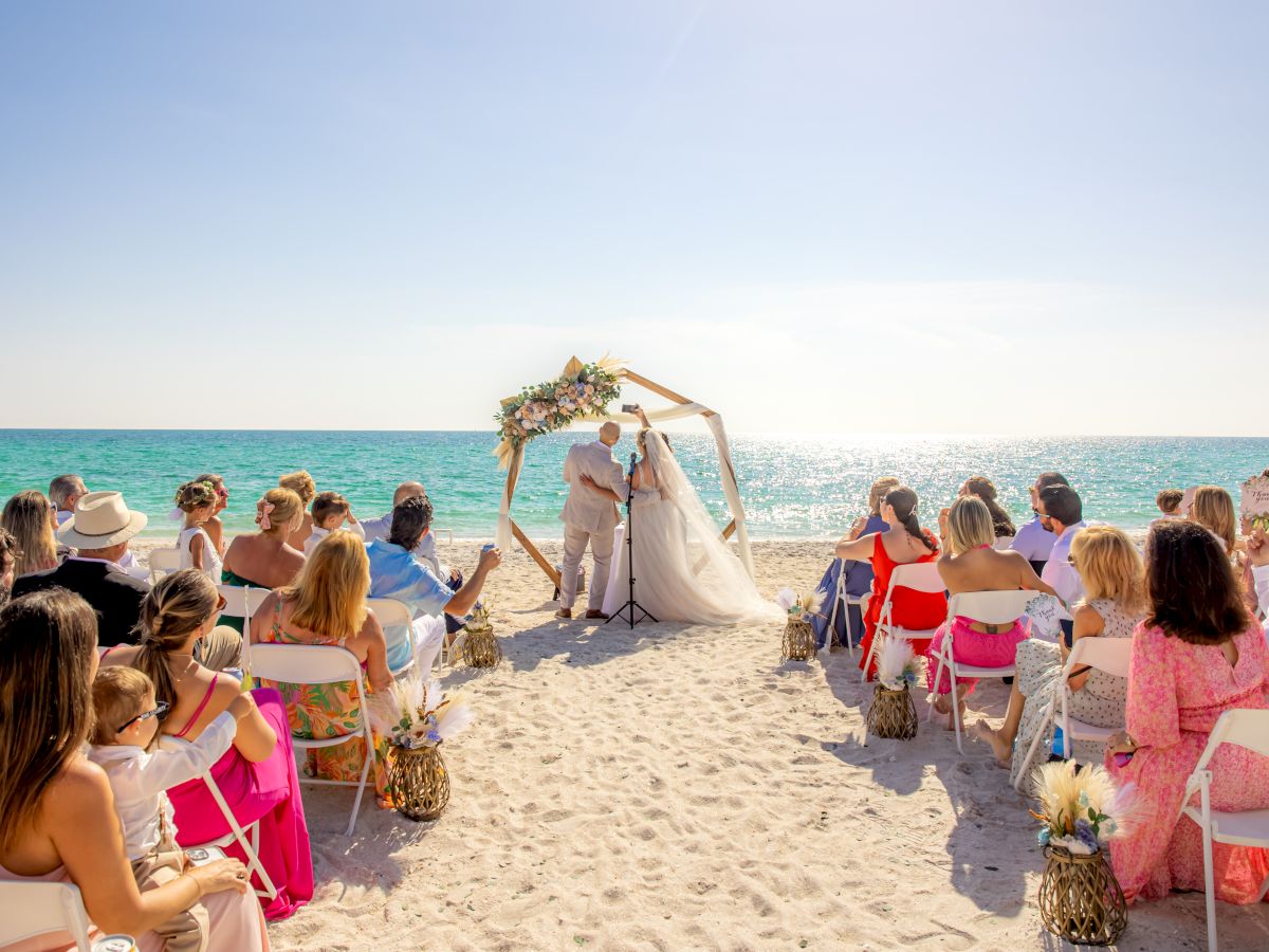 A beach wedding ceremony with guests seated on both sides, the couple at the altar, and an ocean backdrop. Bright, sunny day with clear skies.