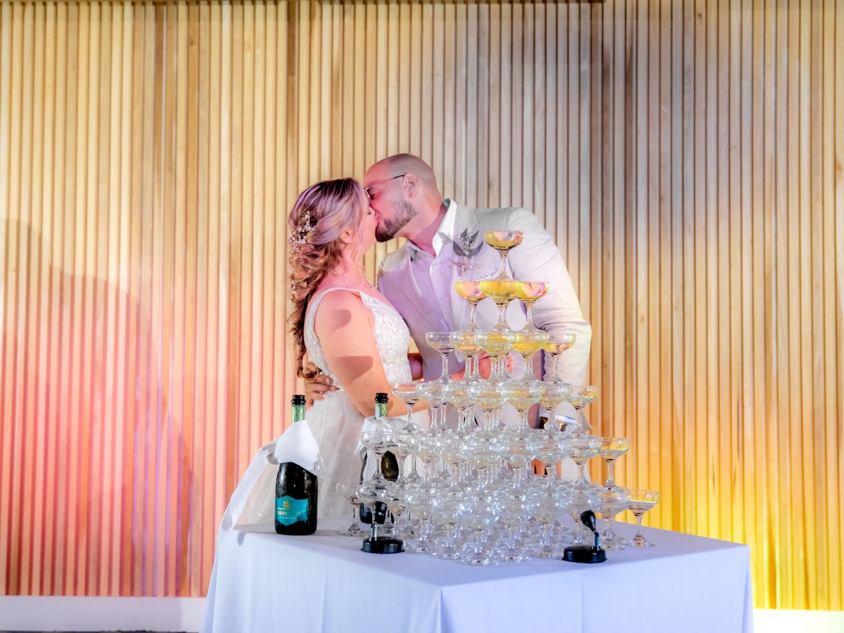 A couple is kissing in front of a champagne tower, with a bottle of champagne on the table covered with a white cloth and a wooden wall backdrop.