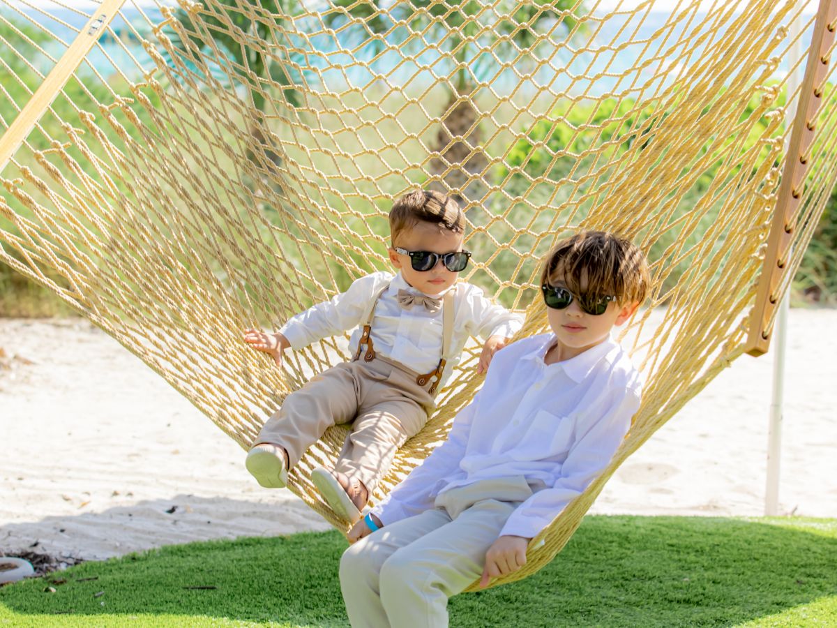 Two children wearing sunglasses are sitting on a hammock in a sunny outdoor setting with grass and palm trees in the background.