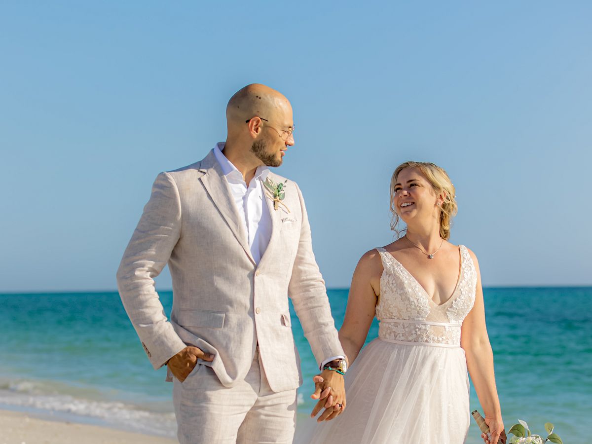 A bride and groom walk hand in hand on a sandy beach by the ocean under a clear blue sky, dressed in wedding attire.