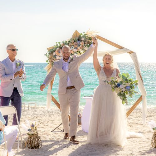 A newlywed couple celebrates on a beach with guests clapping and cheering in the background, set against a scenic ocean view.