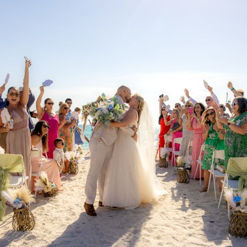 A couple kisses and embraces at a beach wedding ceremony, surrounded by cheering and applauding guests seated on the sand.