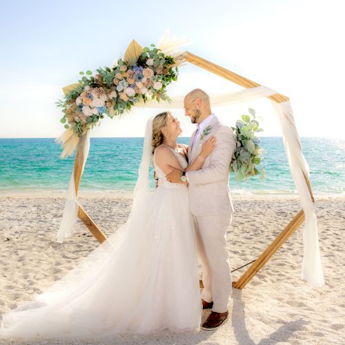 A bride and groom are standing under a floral arch on a beach, with the ocean in the background and the sun shining overhead.