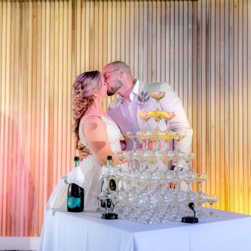 A couple is kissing in front of a champagne tower at a celebration, with a bottle of champagne on a table covered with a white cloth.