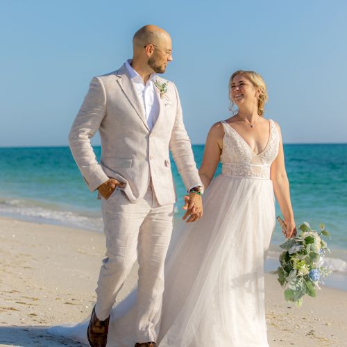 A couple dressed in wedding attire walks hand in hand on a sandy beach, with the ocean and clear blue sky in the background.