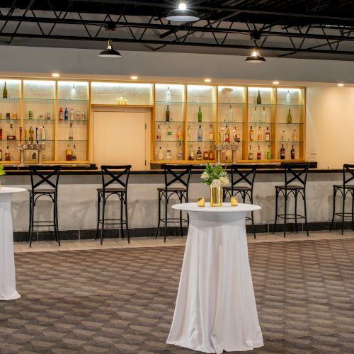 The image shows a modern bar with high chairs, a well-lit display of bottles, and three round tables with white tablecloths and small flower arrangements.