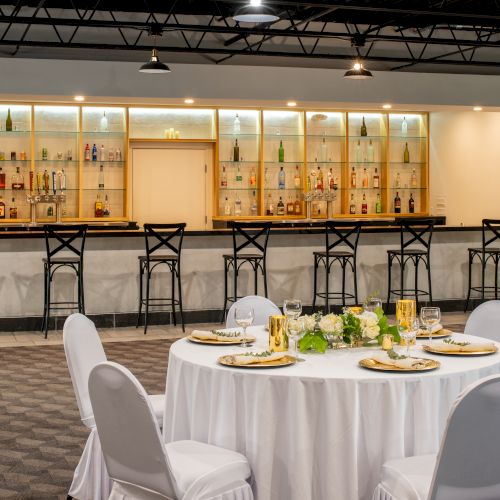 A neatly set table with white linens and elegant tableware is in the foreground, with a stylish well-stocked bar and barstools in the background.