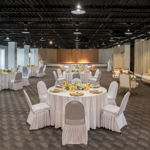 The image shows an elegant banquet hall with round tables covered in white tablecloths, decorated chairs, and a cozy seating area in the back.