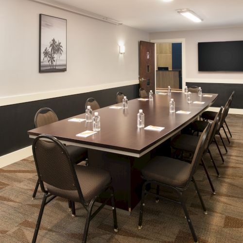 A conference room with a long table, eight chairs, bottled water, and notepads; wall art and a TV screen in the background end the sentence.