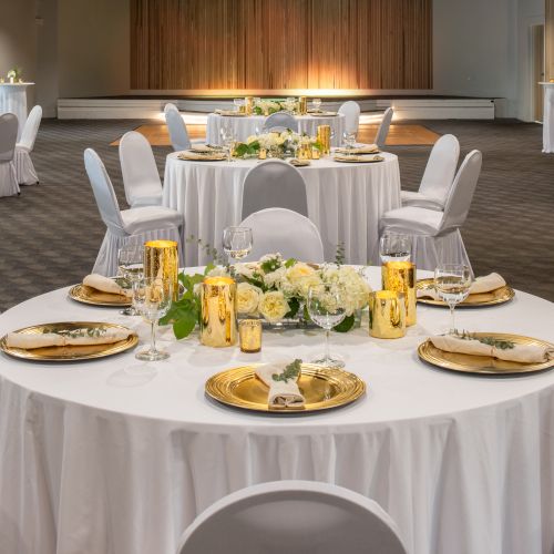 The image shows an elegantly decorated event space with round tables set with white linens, flowers, plates, and golden candle holders.