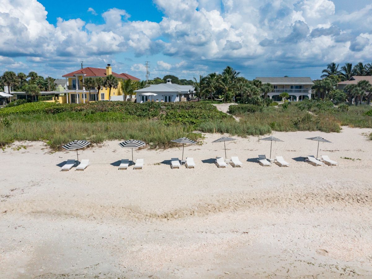 The image shows a beach with empty lounge chairs and umbrellas, with houses and greenery in the background under a partly cloudy sky.