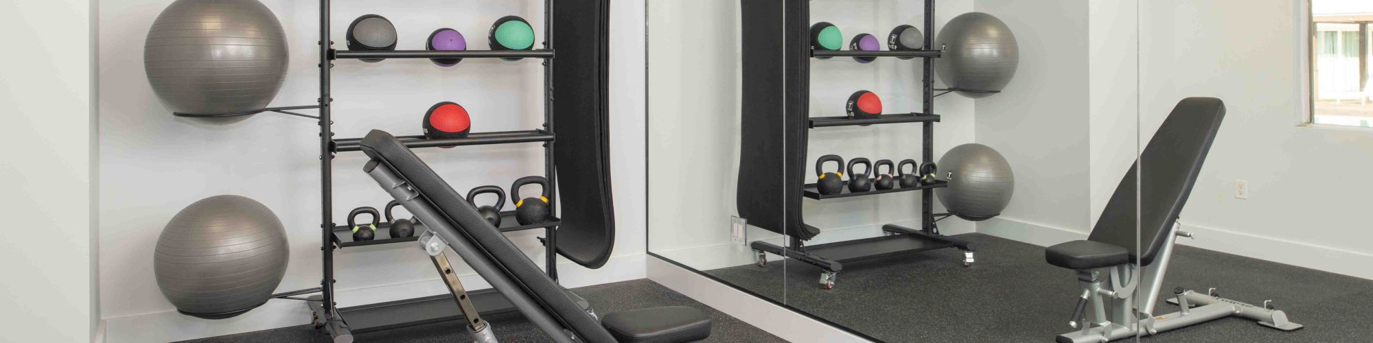 A neatly organized gym space with a bench press, stability balls, kettlebells, medicine balls, and a mirrored wall in a well-lit room.