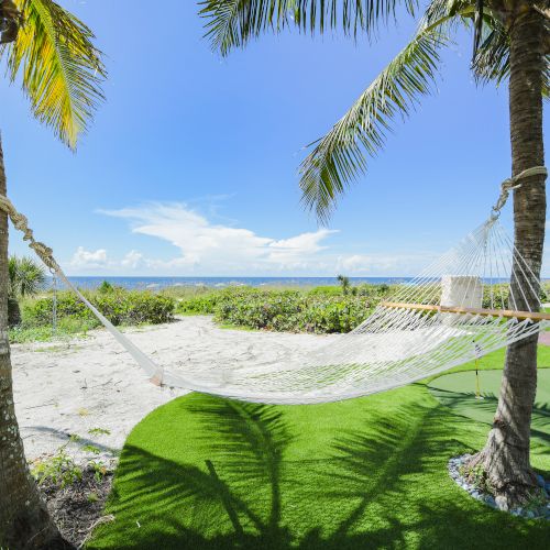 A hammock is strung between two palm trees, overlooking a sandy beach and blue ocean, with lounge chairs and a table nearby.