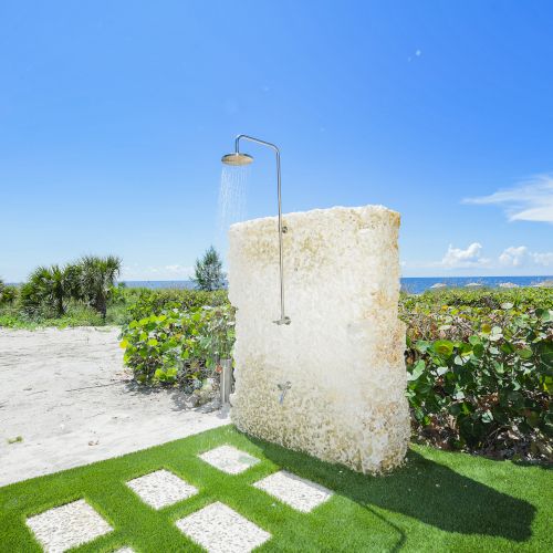 An outdoor shower next to a stone wall, lush greenery, grass pavers, and a scenic view of the ocean under a clear blue sky ending the sentence.