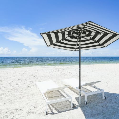 A beach with white sand, two lounge chairs, and a black-and-white striped umbrella overlooking a calm sea under a clear blue sky.