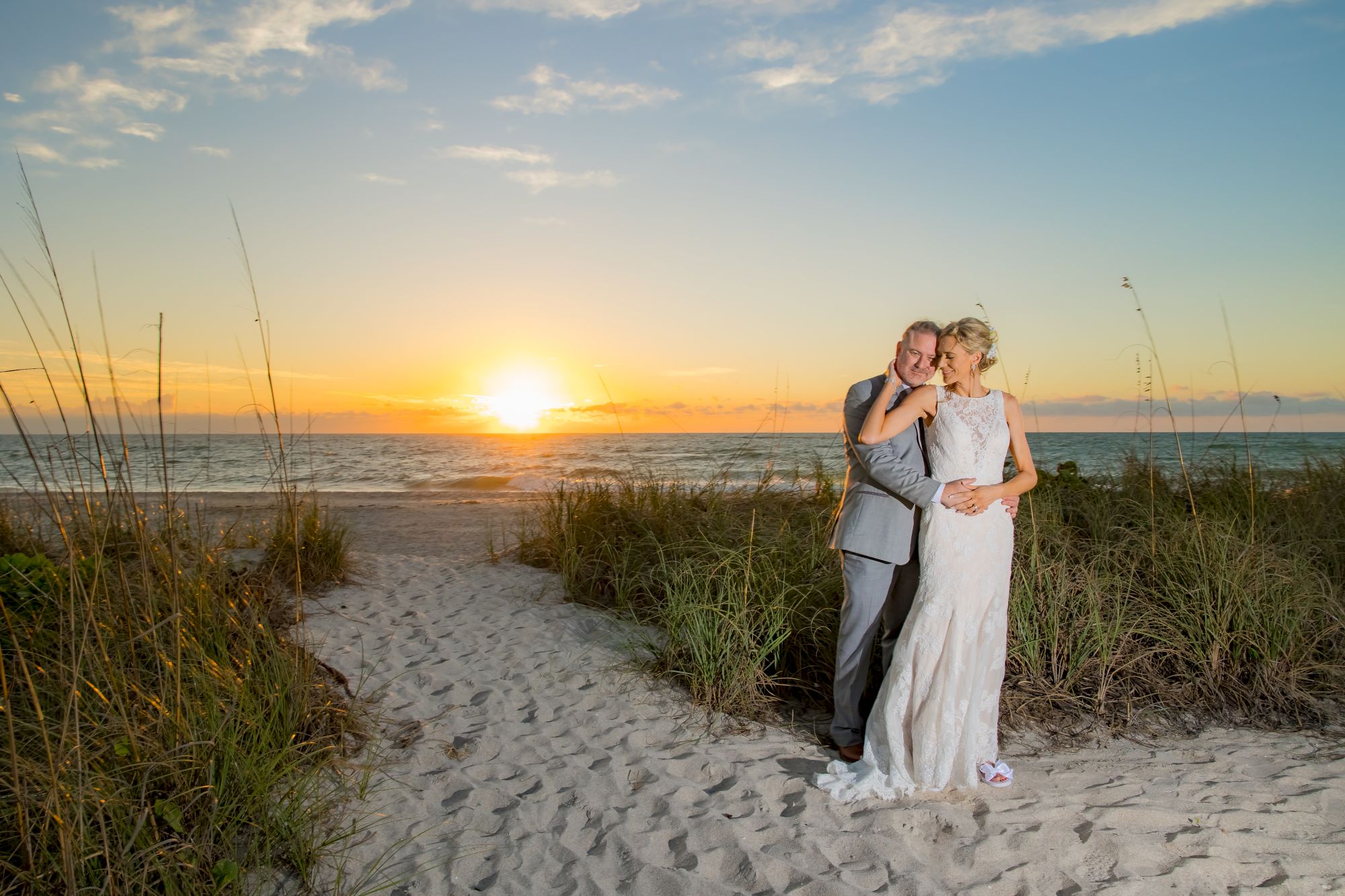 A couple in wedding attire poses on a beach at sunset, surrounded by sand and greenery.