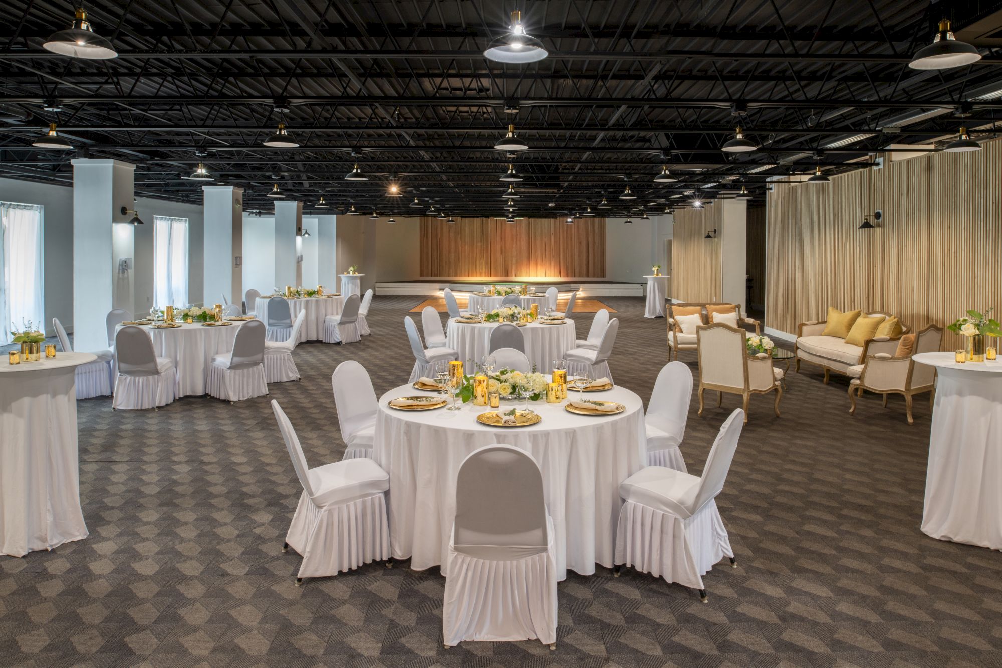 The image shows a decorated event hall with round tables set with white linens and chairs, arranged for a gathering or a banquet, with a stage in the back.