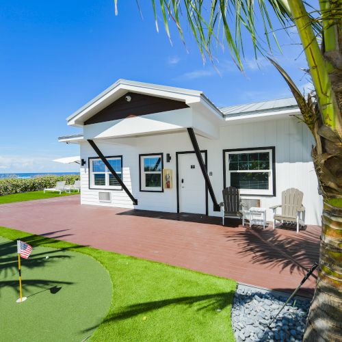 A modern white house with a porch, surrounded by palm trees and a small putting green, under a clear blue sky.