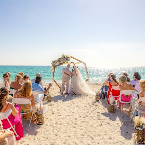 A beach wedding ceremony with guests seated, and a couple under a decorated arch near the ocean on a sunny day.