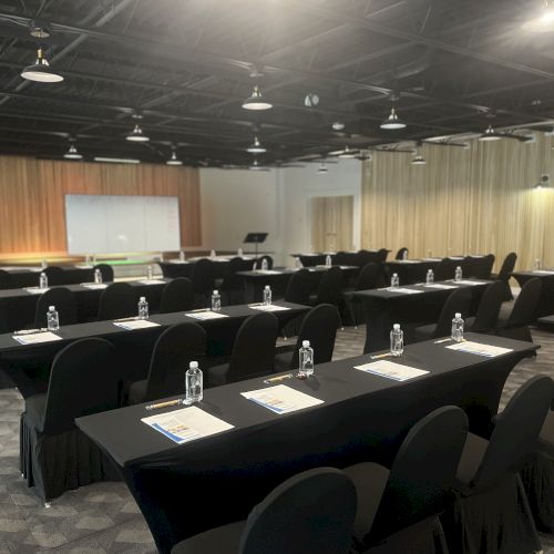 A conference room with rows of tables and chairs, each setup with notepads and water bottles, facing a presentation screen and a podium at the front.