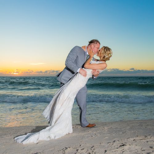 A couple is sharing a kiss on the beach at sunset in wedding attire, with the ocean waves in the background.