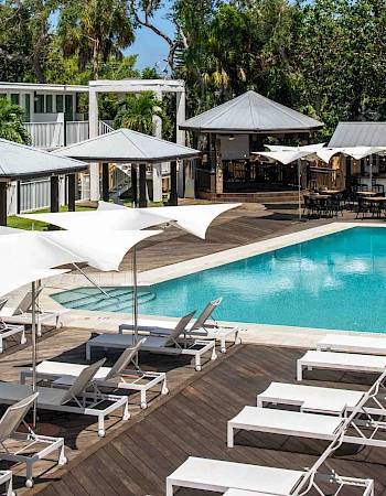 A resort pool area with white lounge chairs, umbrellas, and adjacent buildings set amidst lush greenery and a wooden deck.