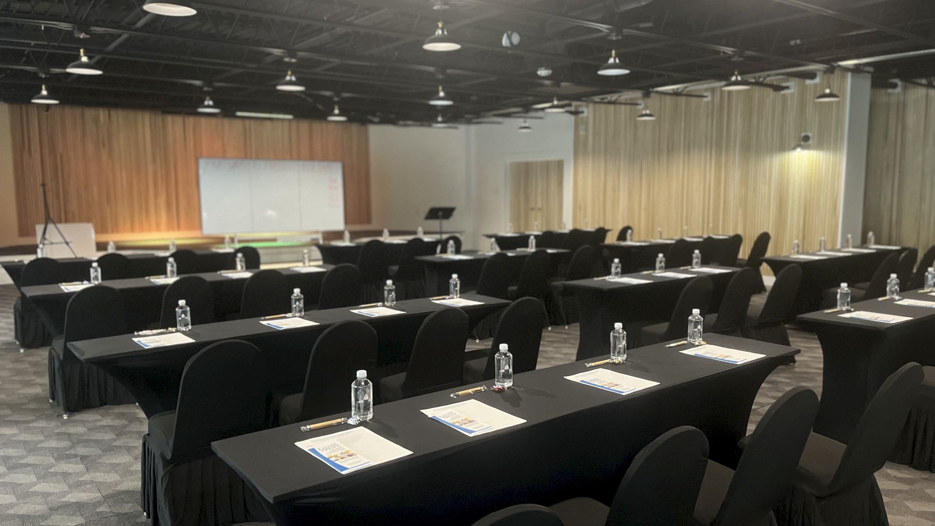 A conference room with rows of tables and chairs, each table set with papers and water bottles, facing a screen and podium.