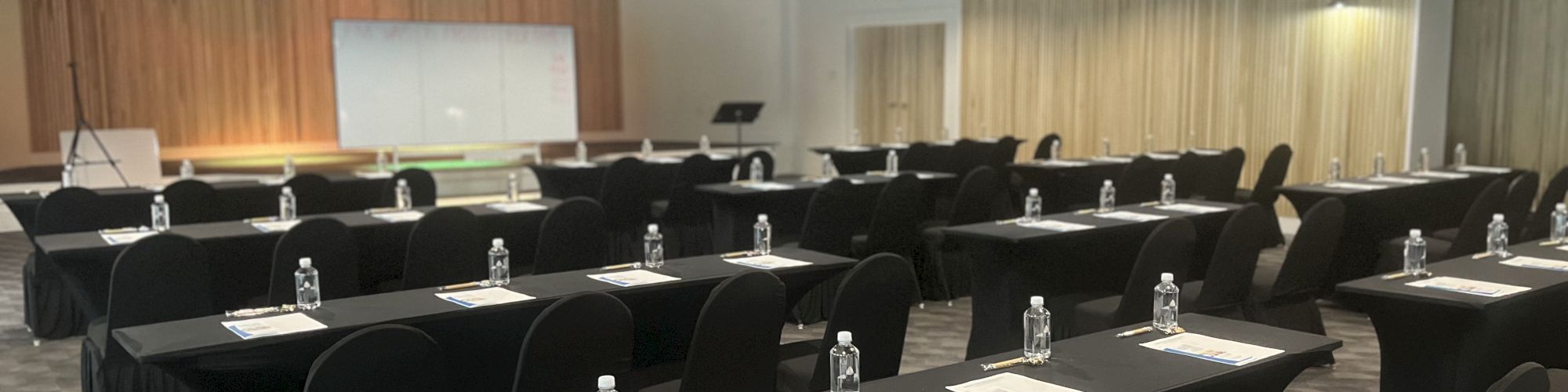 A conference room with rows of tables and chairs, each table set with papers and water bottles, facing a screen and podium.