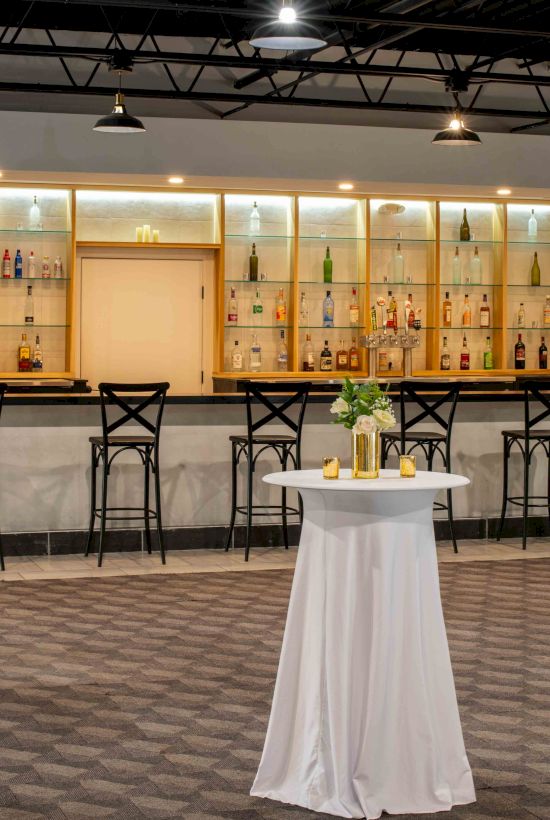 The image shows a modern bar area with high shelves of bottles, barstools, and cocktail tables with white tablecloths and small flower arrangements.