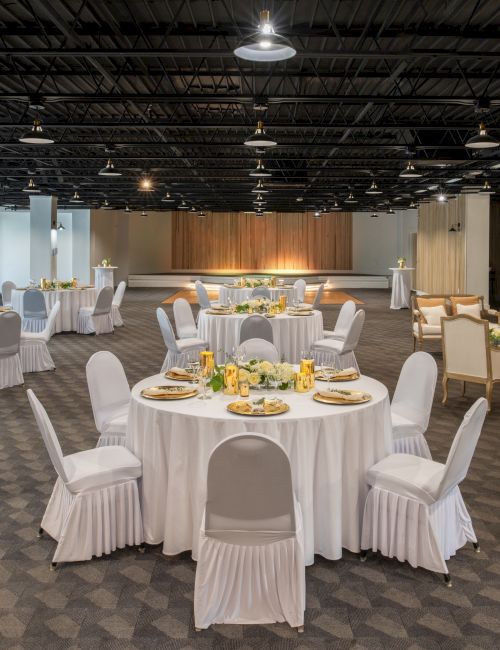 A decorated event space with round tables, white cloths, and chairs; set for a gathering. Sofas are along the wall, under ceiling lights.