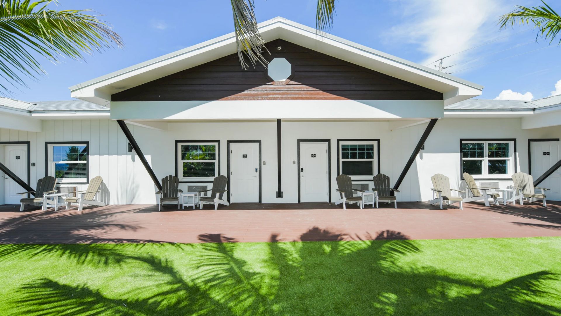 A modern building with white siding and dark accents, featuring multiple doors and windows, and outdoor seating on a sunny day.
