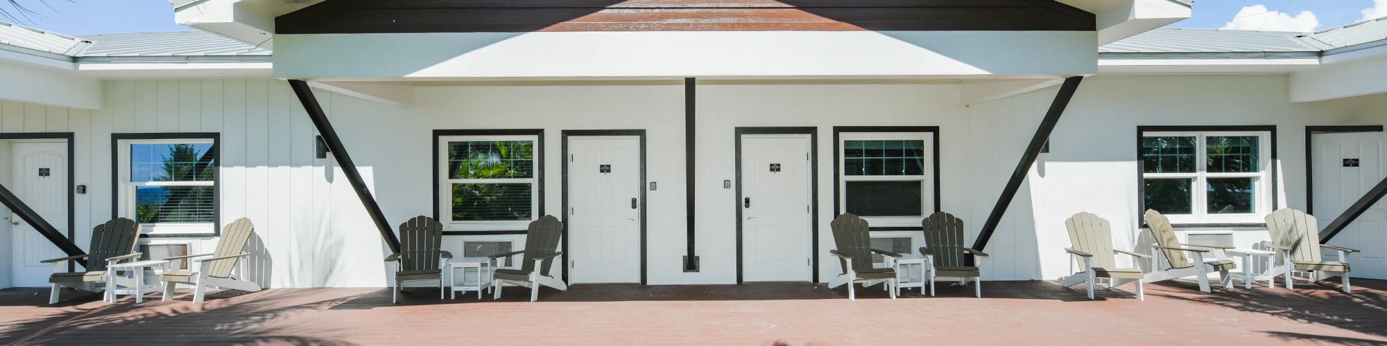 A modern building with white siding and dark accents, featuring multiple doors and windows, and outdoor seating on a sunny day.