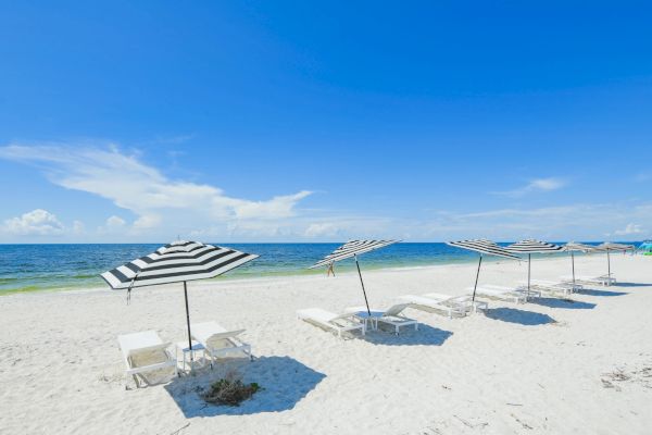 A beach scene with striped umbrellas and lounge chairs on white sand, facing a blue ocean under a clear sky with a few clouds.