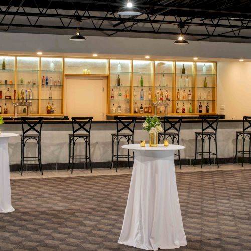 A modern bar area with high tables, white tablecloths, and stools in front of shelves stocked with bottles.