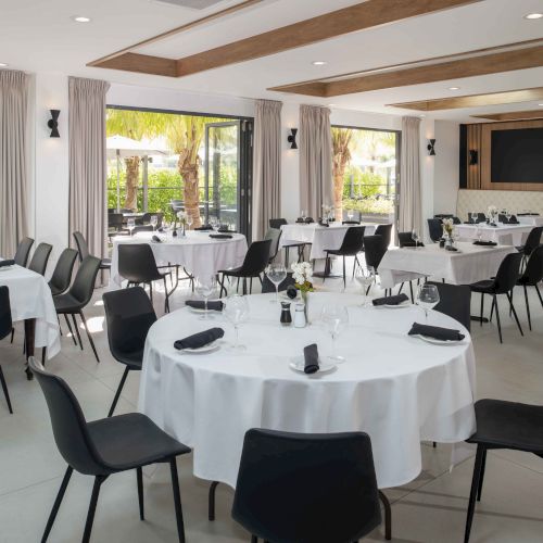 A modern dining room with round tables set with white tablecloths, black chairs, and glassware, with large windows opening to a garden view.