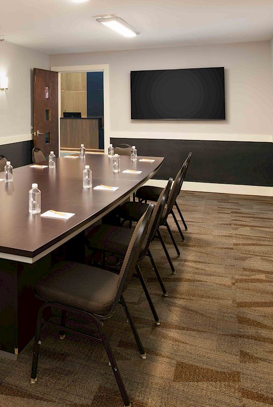 A conference room with a long table, chairs, water bottles, notepads, and a wall-mounted screen is shown.