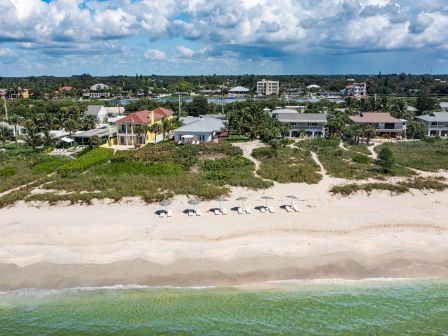 Aerial view of beachfront houses with lush greenery, a sandy beach, and calm ocean waves under a cloudy sky. Beach umbrellas are visible.