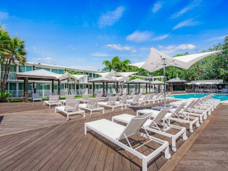 The image shows a poolside area with lounge chairs, umbrellas, wooden deck, palm trees, and a hotel building in the background.