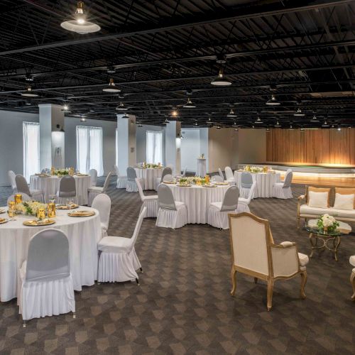 A spacious banquet hall with round tables covered in white cloth, elegant chairs, and a cozy seating area. Soft lighting creates a warm ambiance.